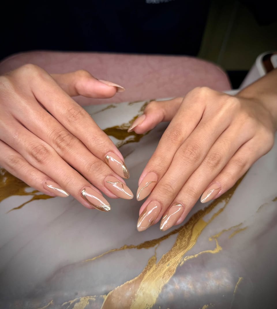 Almond nails in a sheer warm nude with thin, flowing metallic gold lines.