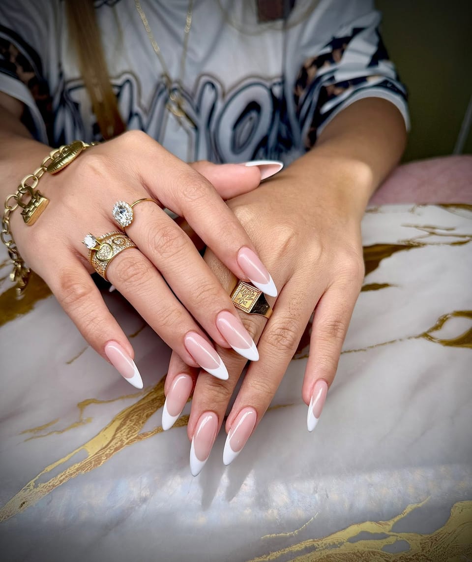 Long almond nails with crisp white French tips on crossed hands wearing gold rings.