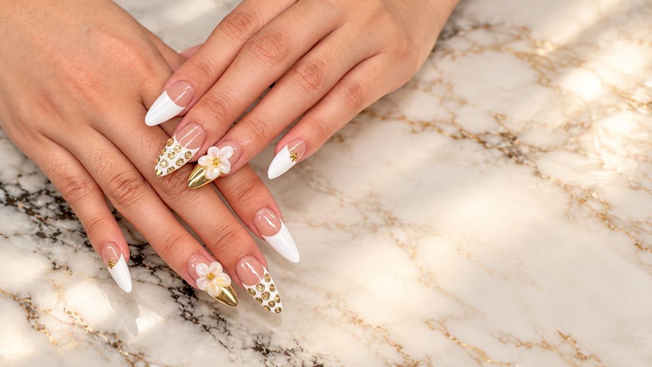 Hands resting on white and gold marble, wearing long almond nails with white French tips, raised cream flowers and gold stud accents.